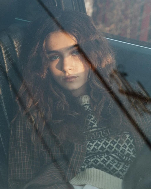 Portrait of a young woman with wavy brown hair in an automotive interior, wearing a patterned sweater and jacket, gazing at the camera.