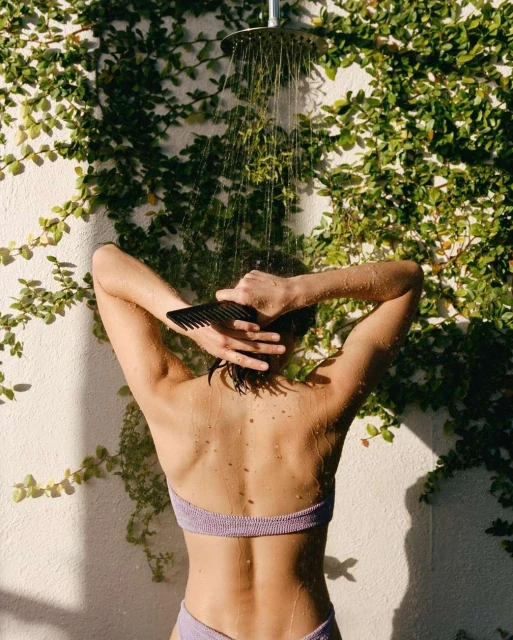 Woman showering outdoors surrounded by ivy. She is facing away, combing her hair with a black comb under the showerhead.