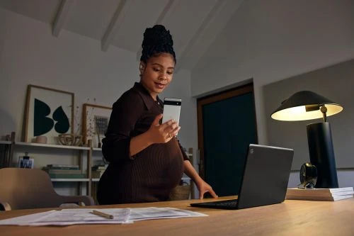 Pregnant woman in brown dress standing at desk, holding phone, with laptop, lamp, and artwork in background at home during the day.