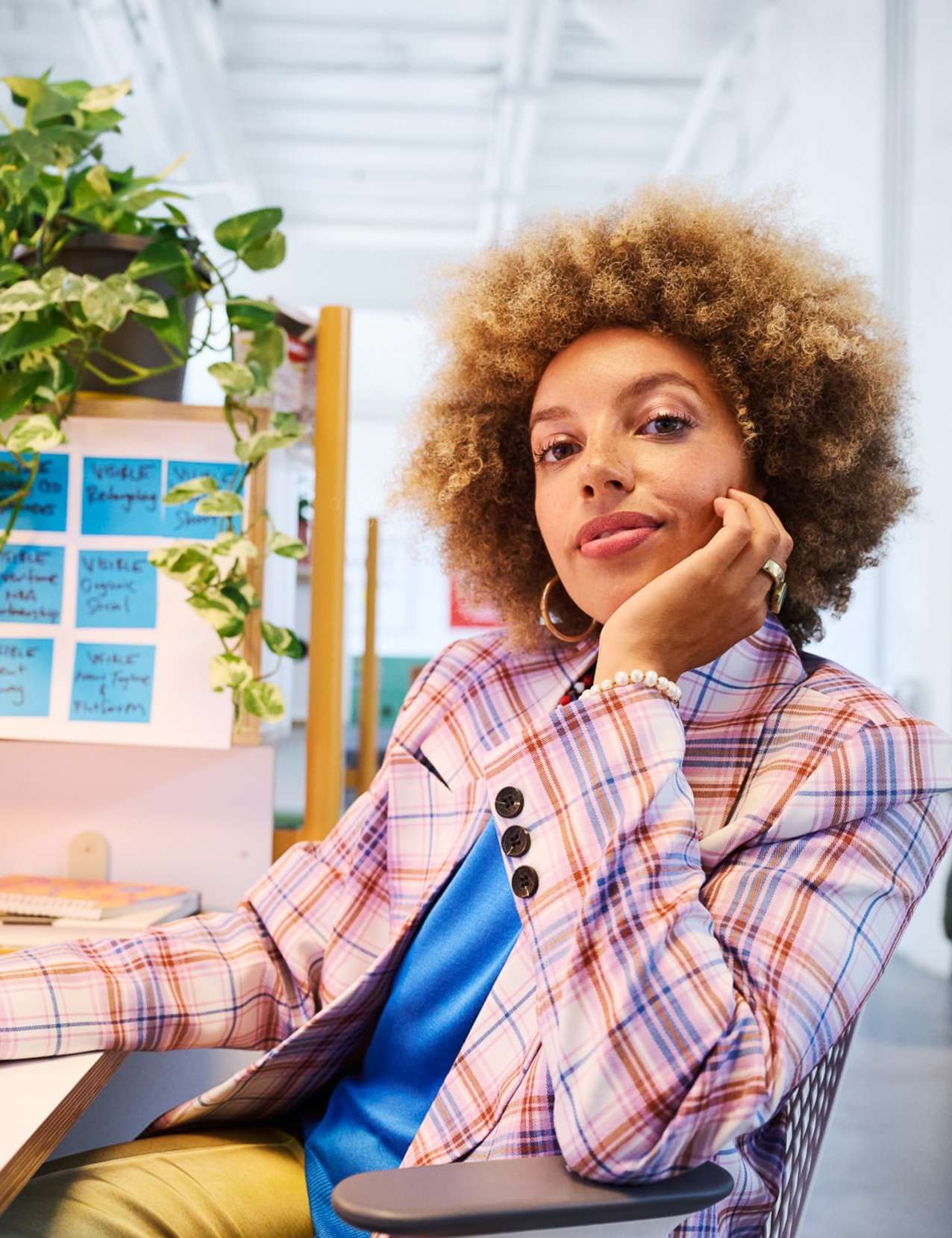 Portrait of a stylish woman with curly hair wearing a plaid blazer, posing indoors in an office setting with plants in the background.