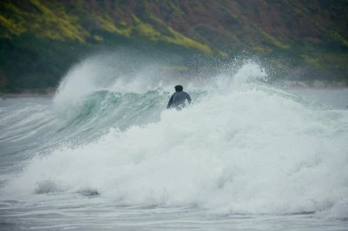 A person is surfing a large wave, with water splashing all around them, on a cloudy day with a green hill in the background.