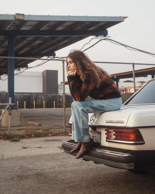 A brunette woman sits on the trunk of a vintage white Mercedes car outside on a cloudy day, gazing off into the distance.
