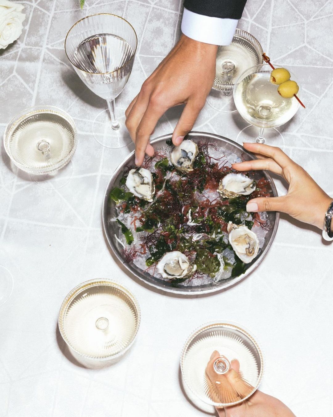 Overhead shot of an oyster platter surrounded by cocktails on a white linen tablecloth, hands of two adults are visible.