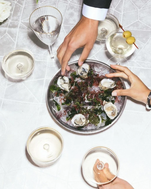 Overhead shot of an oyster platter surrounded by cocktails on a white linen tablecloth, hands of two adults are visible.