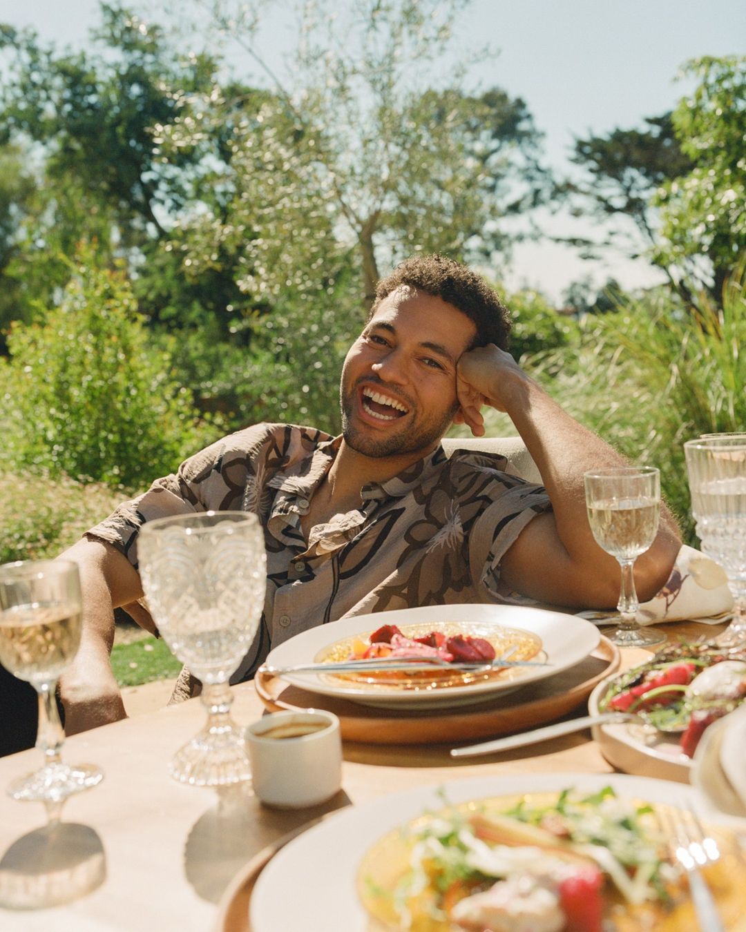 Smiling man seated at an outdoor dining table set with plates of food and wine glasses under trees