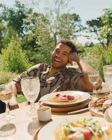 Smiling man seated at an outdoor dining table set with plates of food and wine glasses under trees