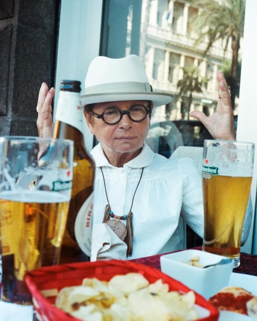 Woman in hat with glasses raises her hands at an outdoor cafe, sitting behind two beer bottles and a bowl of chips, city buildings behind.