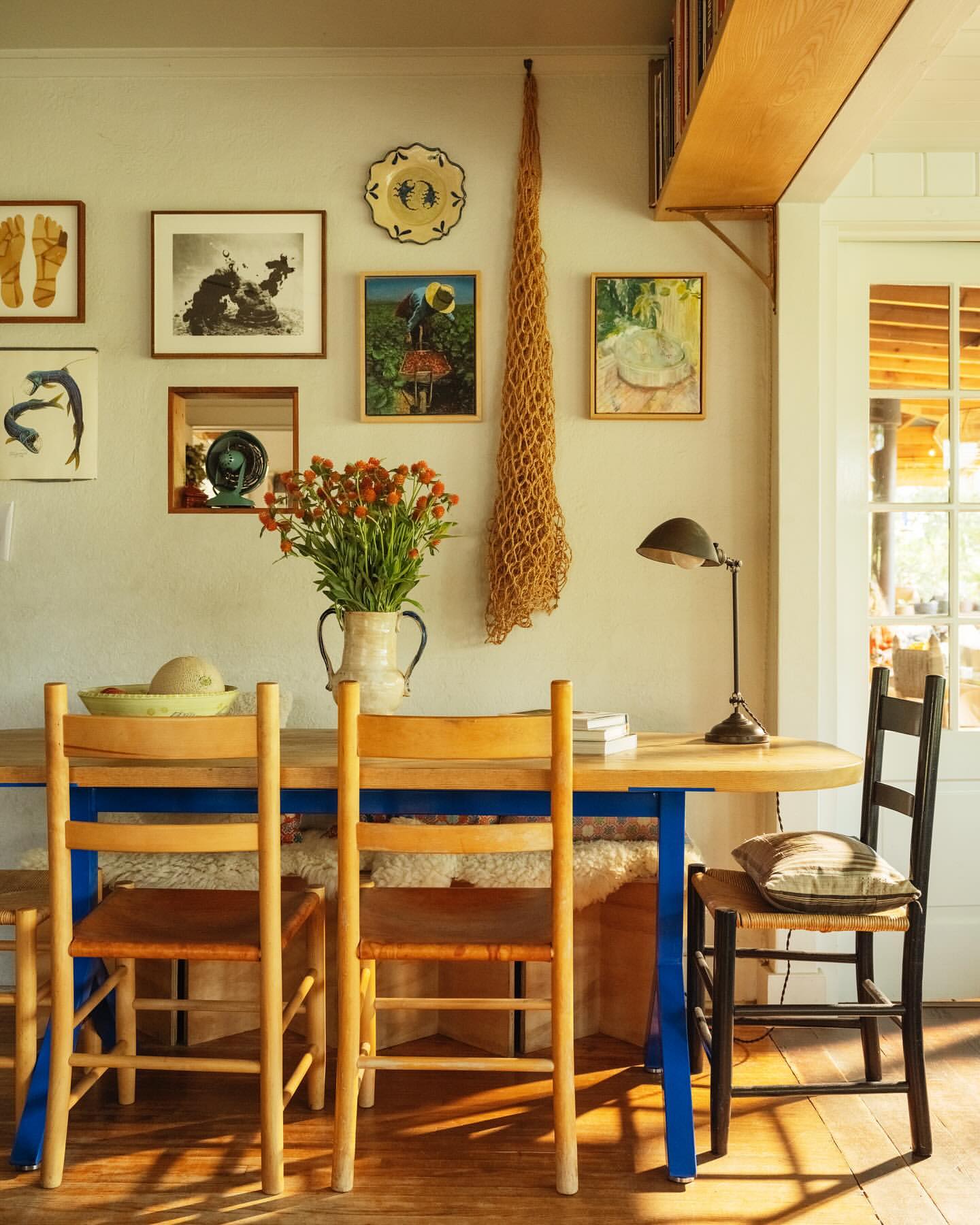 Still life of a rustic dining room with a wooden table, woven chairs, and orange flowers in a ceramic pitcher against a gallery wall.