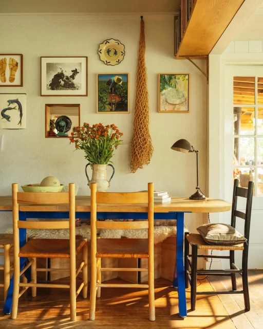 Still life of a rustic dining room with a wooden table, woven chairs, and orange flowers in a ceramic pitcher against a gallery wall.