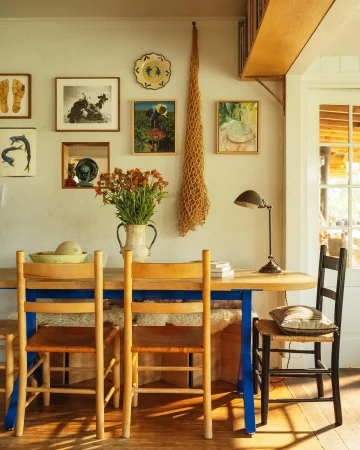 Still life of a rustic dining room with a wooden table, woven chairs, and orange flowers in a ceramic pitcher against a gallery wall.