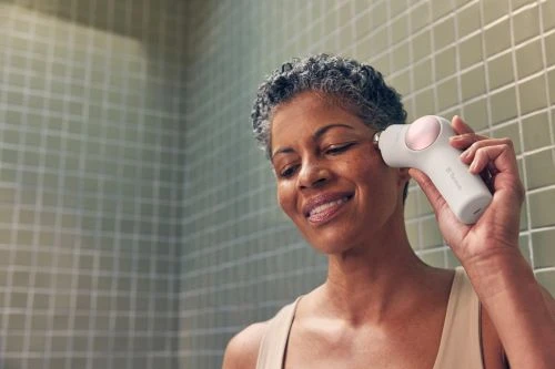 An adult woman uses a facial toning device in front of a tiled wall, showing skincare routine and beauty technology for skin rejuvenation.
