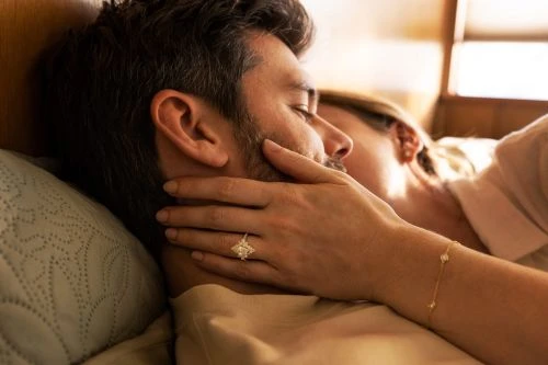 Close-up of a man and woman couple sleeping in bed, the woman's hand resting on the man's face with a diamond ring visible.