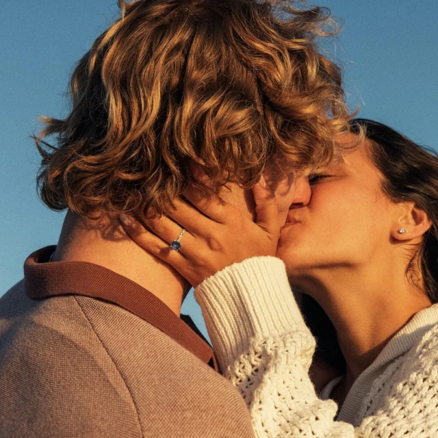 Close-up portrait of a kissing couple in soft light, featuring a woman with brown hair and a man with blonde hair, against a blue sky background.