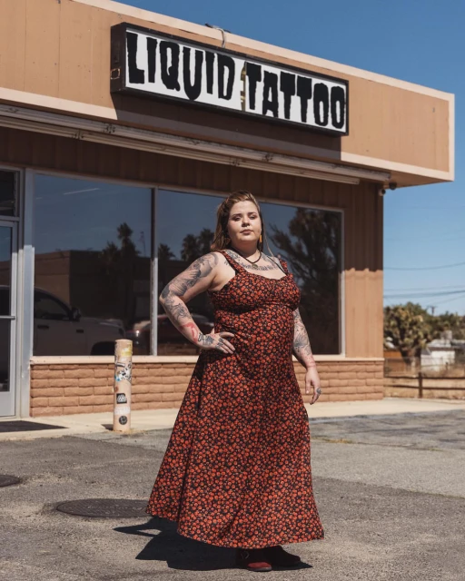 Adult woman with tattoos stands in a red floral dress outside Liquid Tattoo, a single-story building under a clear sky in daytime.