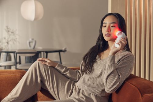 Asian woman uses a red light therapy device on her face while sitting on an orange couch in a well-lit interior space.