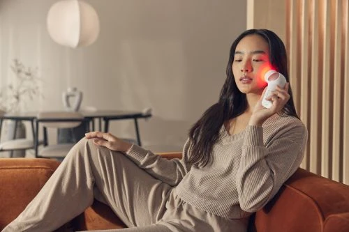 Asian woman uses a red light therapy device on her face while sitting on an orange couch in a well-lit interior space.