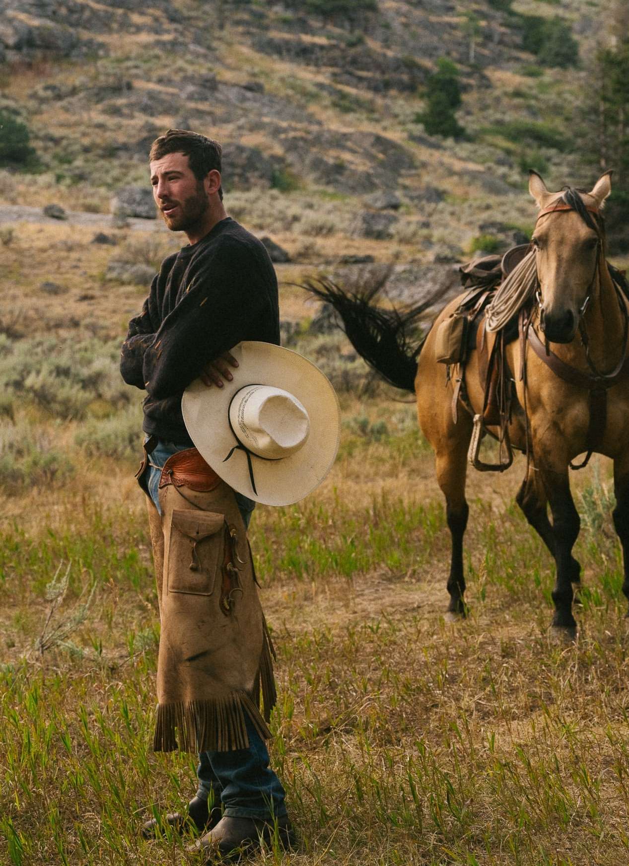Man standing in a field with a horse and a hillside in the background, wearing chaps and holding a cowboy hat.