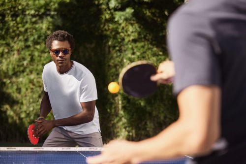 Two men playing ping pong outdoors in natural light, one in focus with sunglasses and a white t-shirt, green trees in background.