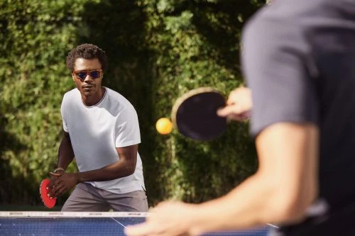 Two men playing ping pong outdoors in natural light, one in focus with sunglasses and a white t-shirt, green trees in background.