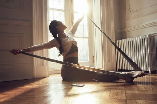 Adult female performs seated stretching exercise with a resistance band indoors, with natural light streaming through a window.
