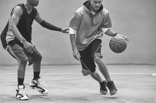 Two adult male basketball players in athletic wear compete on an outdoor court, focusing on dribbling and defense in monochrome.