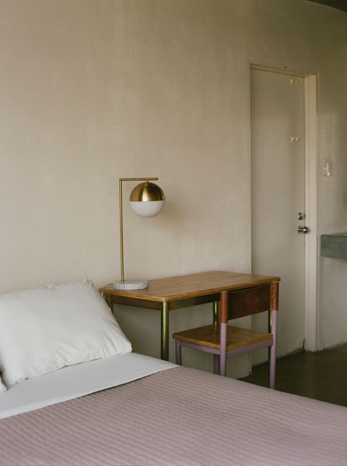 Minimalist hotel bedroom with a neatly made bed, desk, chair, and gold lamp against a light beige wall and white door.