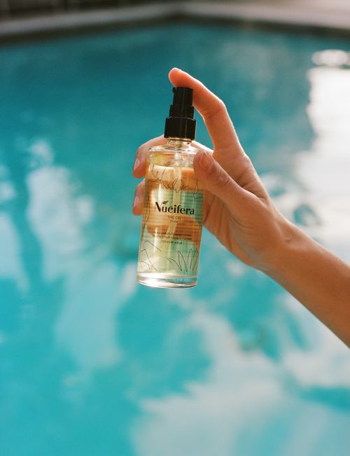 Close-up of a hand holding a bottle of Nueifera product in front of a blurred blue pool, showcasing beauty and wellness.