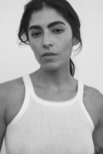 Black and white portrait of a young adult woman with dark hair in a white tank top against a neutral background, facing the camera.