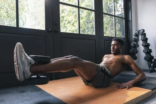 An athletic, shirtless man is doing ab exercises with a dumbbell between his feet on a tan exercise mat indoors with natural light.