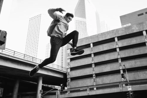 A man leaps across an urban concrete structure against a backdrop of city skyscrapers in a dynamic black and white action shot.