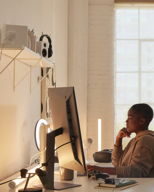 Woman in beige hoodie seated at desk with computer, VR headset, gaming controller, ring light, against white walls and window light.