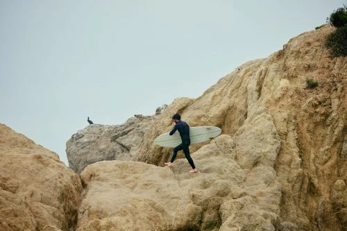 A surfer in a wetsuit carries a surfboard while climbing on rocks, with a bird perched on a rock in the background against a cloudy sky.