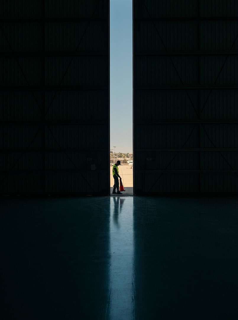 Airport worker in high-visibility clothing stands in a large hangar doorway, holding a safety cone, with a blurred airport in background.