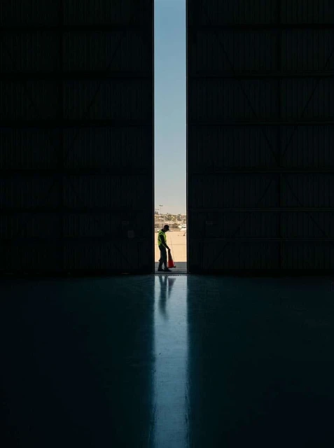 Airport worker in high-visibility clothing stands in a large hangar doorway, holding a safety cone, with a blurred airport in background.