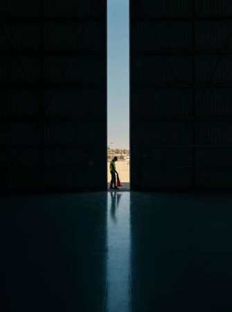 Airport worker in high-visibility clothing stands in a large hangar doorway, holding a safety cone, with a blurred airport in background.