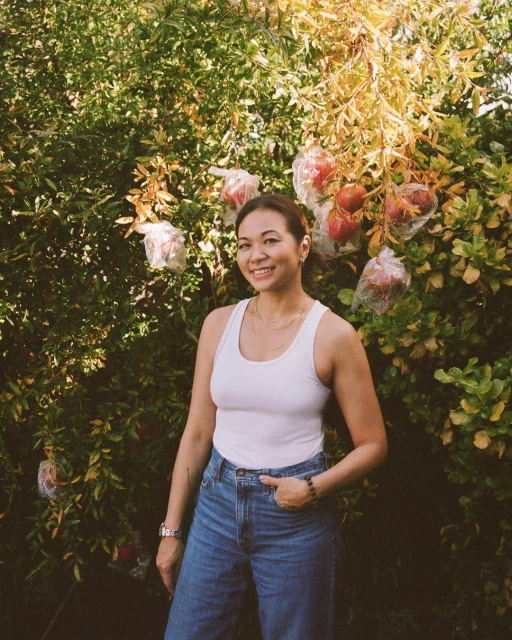 Smiling Asian woman in a white tank top and jeans stands outdoors in front of a leafy tree with red fruit covered in plastic bags.