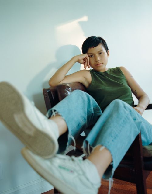 A young woman with short hair poses in an indoor setting wearing a green tank top and light blue jeans while seated in a chair.