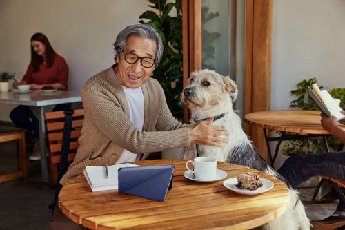 Elderly man pets a dog at an outdoor cafe. A notebook, tablet, coffee, and pastry are on the table. Two people are in the background.