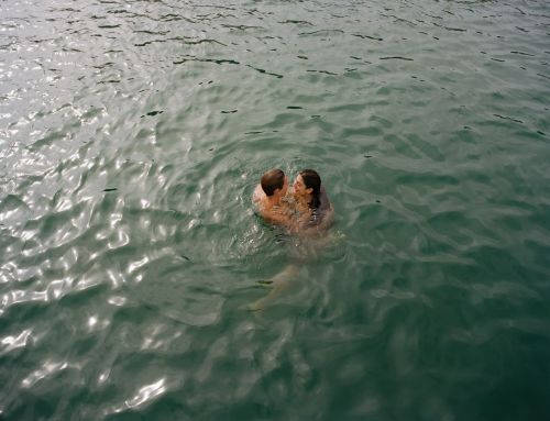 A couple embraces in green water, creating ripples; a sunny day scene with a sense of intimacy and connection in a natural setting.