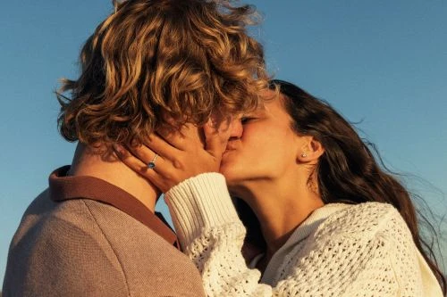 Close-up of a young adult couple kissing outdoors in the sunlight. The woman wears a white sweater and the man wears a brown polo shirt.