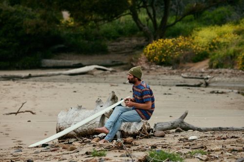 A man in a green beanie and striped shirt sitting on a log on a sandy beach next to a surfboard