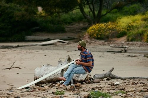 A man in a green beanie and striped shirt sitting on a log on a sandy beach next to a surfboard