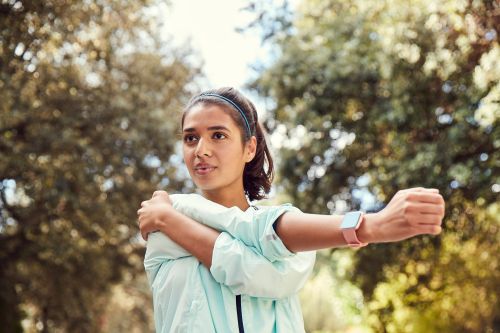 Adult woman in workout attire stretches arm in a park environment with green trees and blue sky. She is wearing a smart watch.