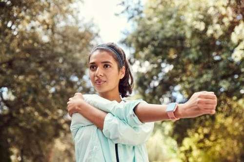 Adult woman in workout attire stretches arm in a park environment with green trees and blue sky. She is wearing a smart watch.