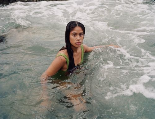 A woman in a green bathing suit sits in wavy ocean water, looking directly at the viewer with a neutral expression on a sunny day.