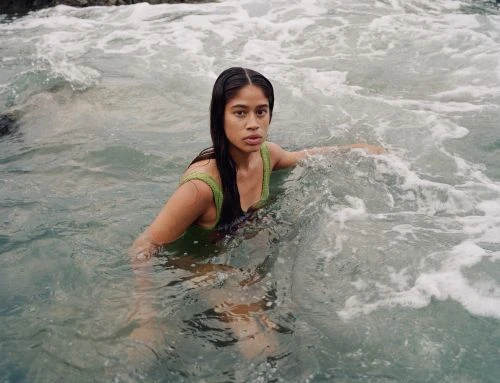 A woman in a green bathing suit sits in wavy ocean water, looking directly at the viewer with a neutral expression on a sunny day.