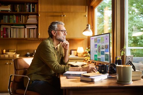 A man wearing glasses works at a computer on a wooden desk in a home office with a bookcase and a large window providing natural light.