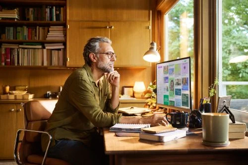 A man wearing glasses works at a computer on a wooden desk in a home office with a bookcase and a large window providing natural light.