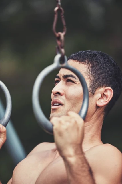 Muscular man working out on gymnastic rings outdoors in a close-up shot against blurred background. Fitness lifestyle.