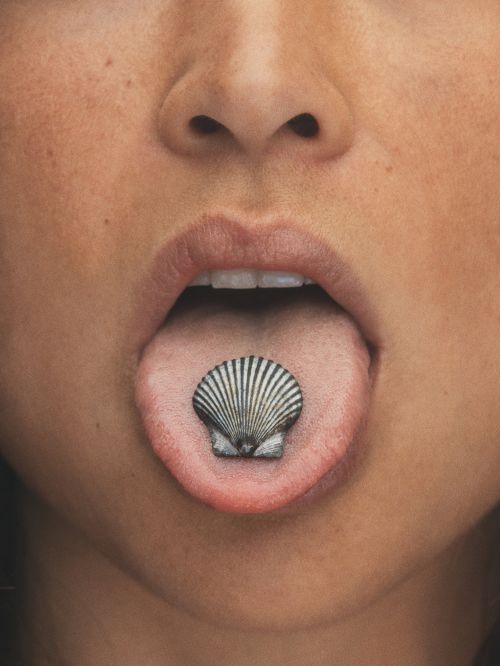 Close-up of a woman's mouth with a seashell on her tongue, set against a blurred background, beauty, body modifications, and fashion.